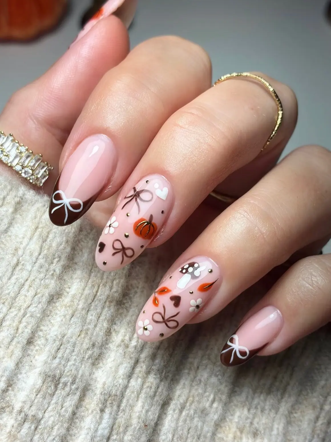 Autumn nail art with a tiny pumpkin, mushrooms, leaves and brown bow French tips against a knit sweater