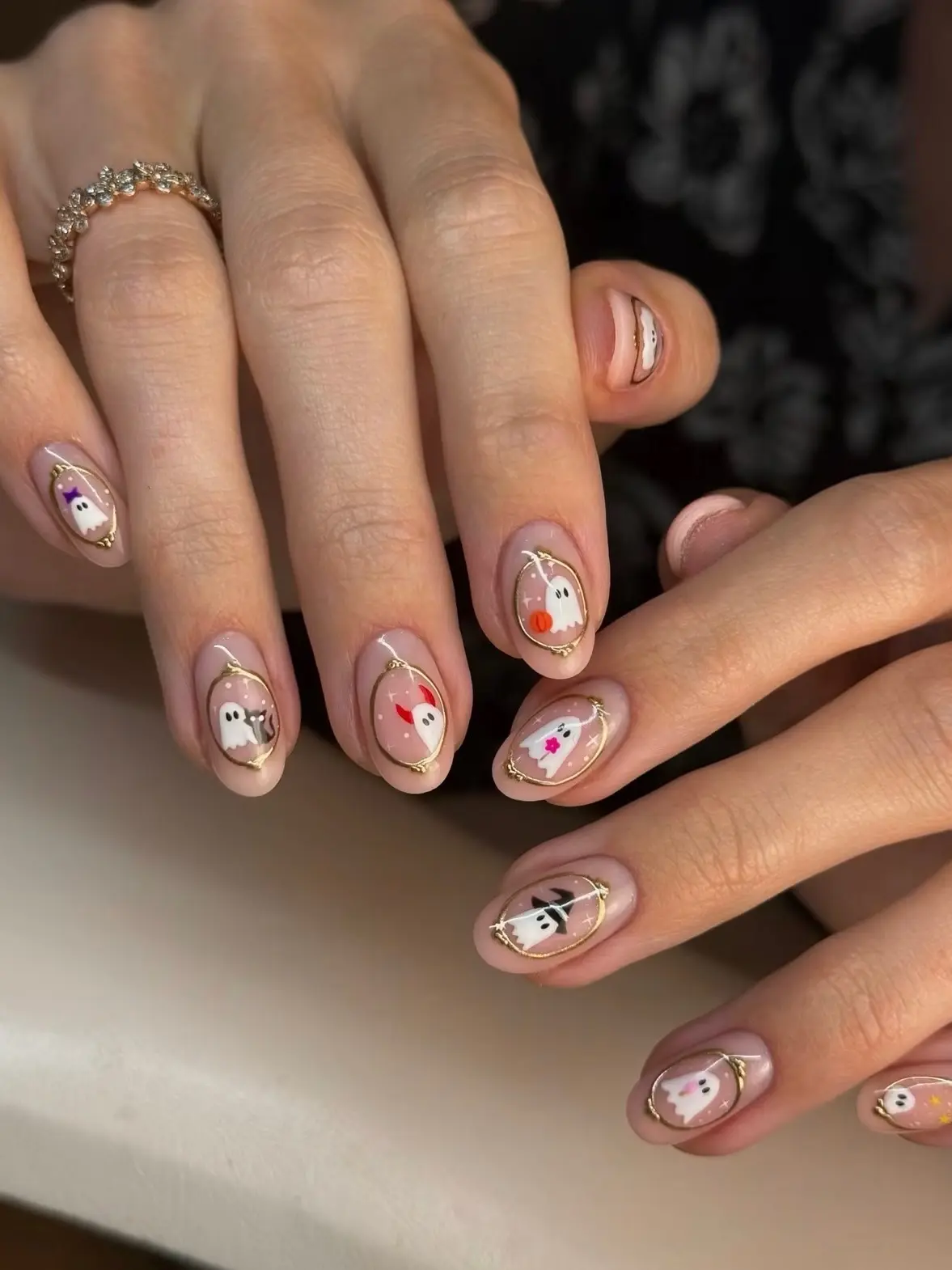Short round Halloween nails with tiny painted ghosts in gold oval frames over a sheer nude base