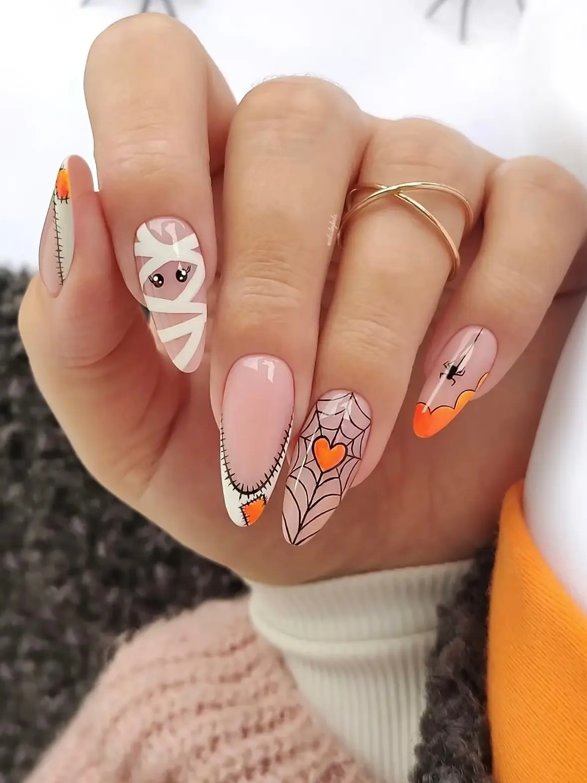 Long almond nails with a mummy face, an orange spiderweb heart, stitched white tips and a hanging spider